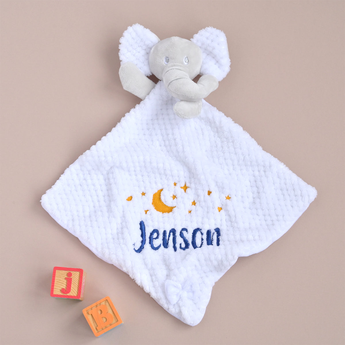 A white waffle-textured baby security blanket with a plush elephant head handle, personalized with the name 'Jenson' in blue and embroidered yellow crescent moon and stars, displayed on a beige background with toy blocks.