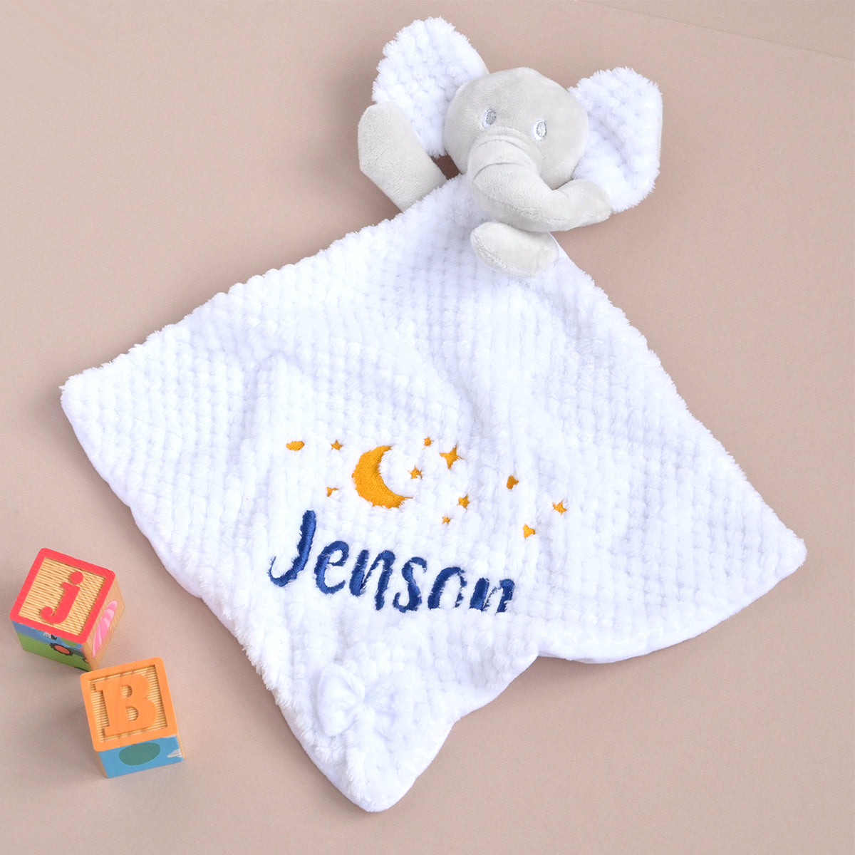 A white waffle-weave baby security blanket with a plush elephant head and embroidered name 'Jenson' in blue with yellow stars and a crescent moon, displayed on a beige background with toy blocks.