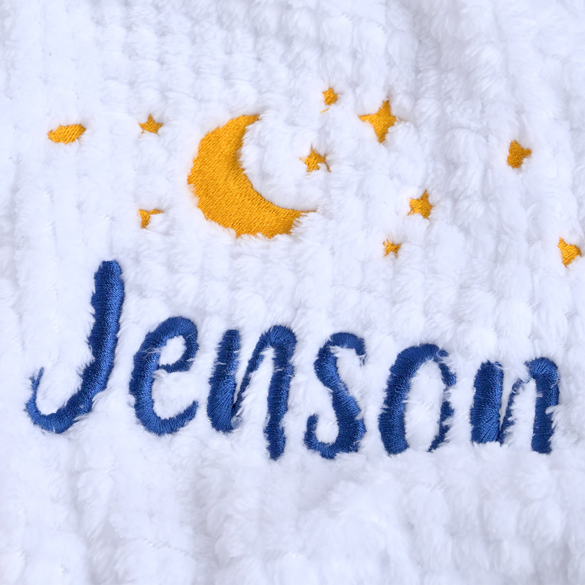 Close-up of a white textured blanket featuring embroidered blue cursive text reading 'Jenson' and a yellow crescent moon with matching yellow stars.