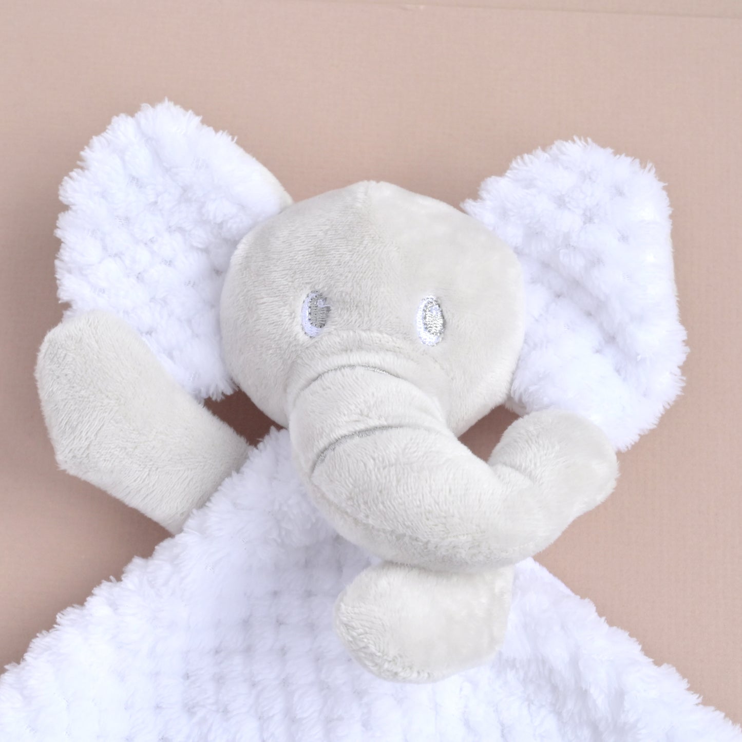 A plush elephant comforter featuring a light gray elephant head and trunk with fluffy white textured ears and blanket, shown against a solid beige background.