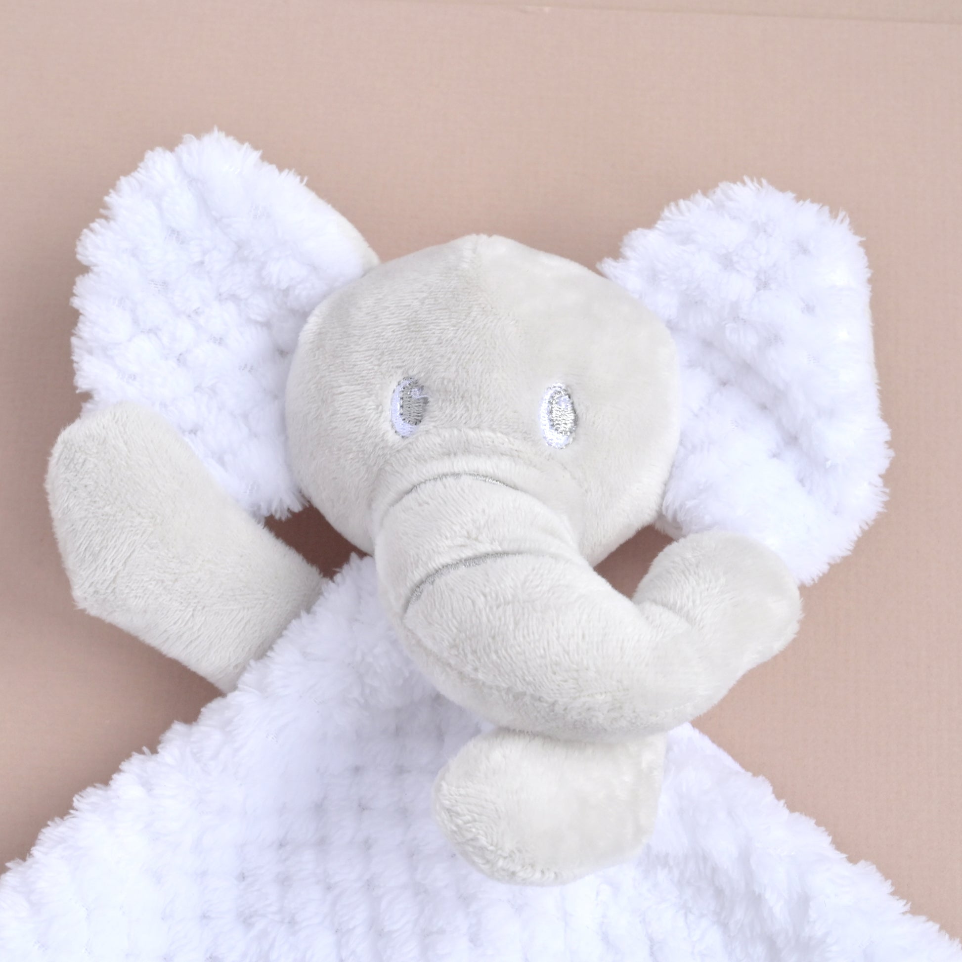 A plush elephant comforter featuring a light gray elephant head and trunk with fluffy white textured ears and blanket, shown against a solid beige background.