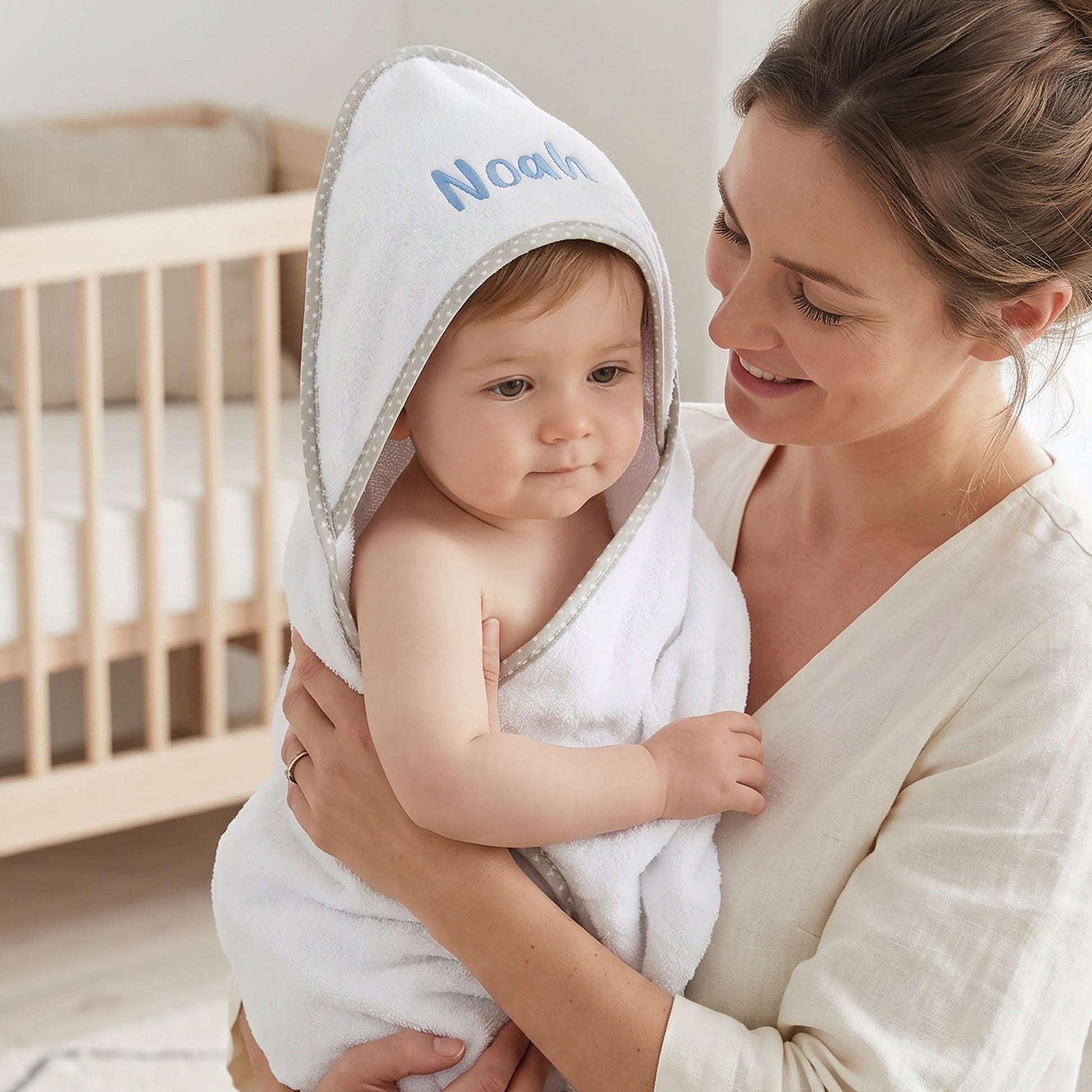 White hooded baby towel with a grey star-print trim around the hood, personalised with the name Noah in blue, worn by a baby wrapped in a mother's arms.