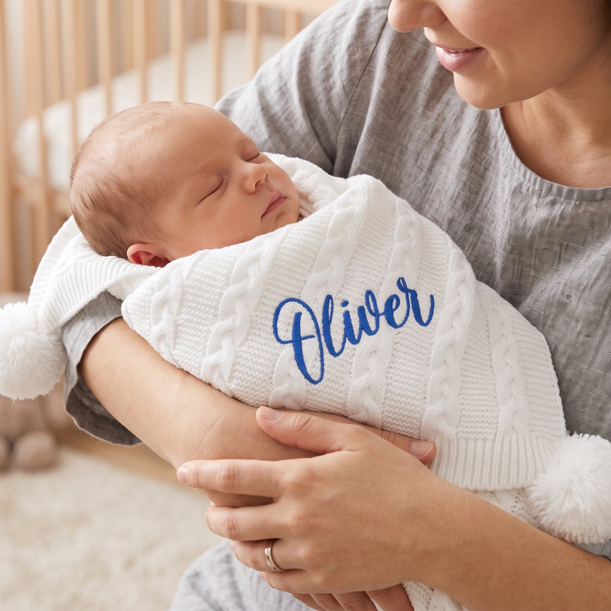 A newborn baby wrapped in a white cable-knit blanket with fluffy pom-poms and the name 'Oliver' embroidered in blue script, held by a mother wearing a gray shirt.