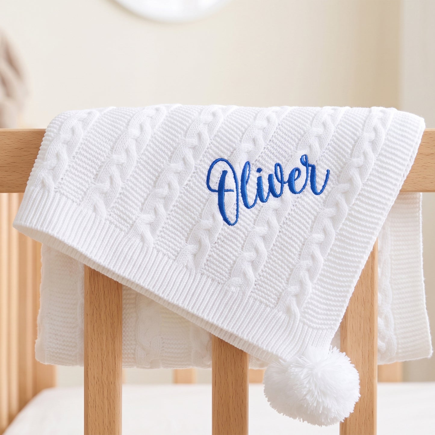 A white cable-knit baby blanket with a pom-pom and personalized blue embroidery reading 'Oliver', draped over a light wood crib.