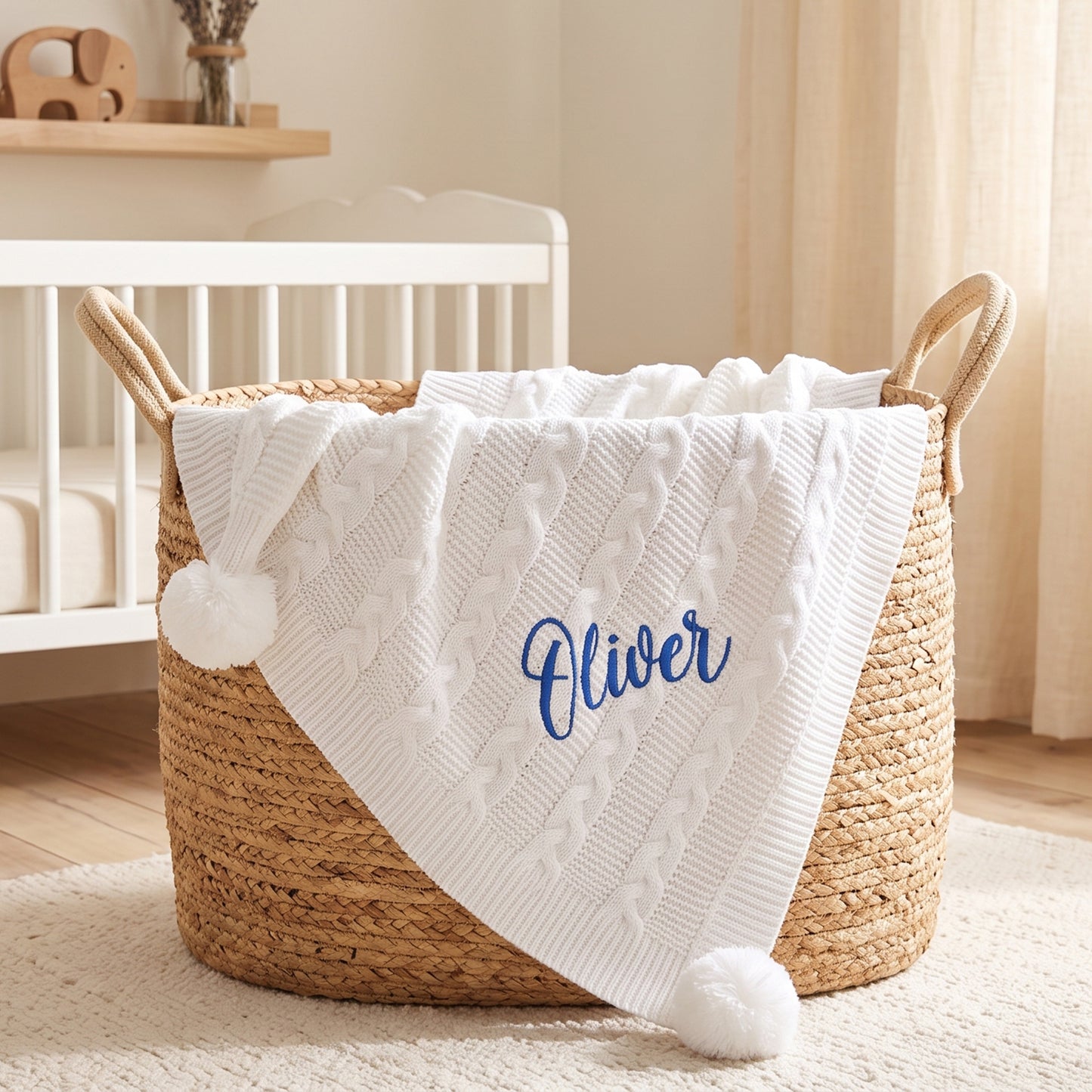 White cable knit baby blanket with pom-poms at each corner, personalised with the name Oliver in blue embroidery, draped over a woven basket.