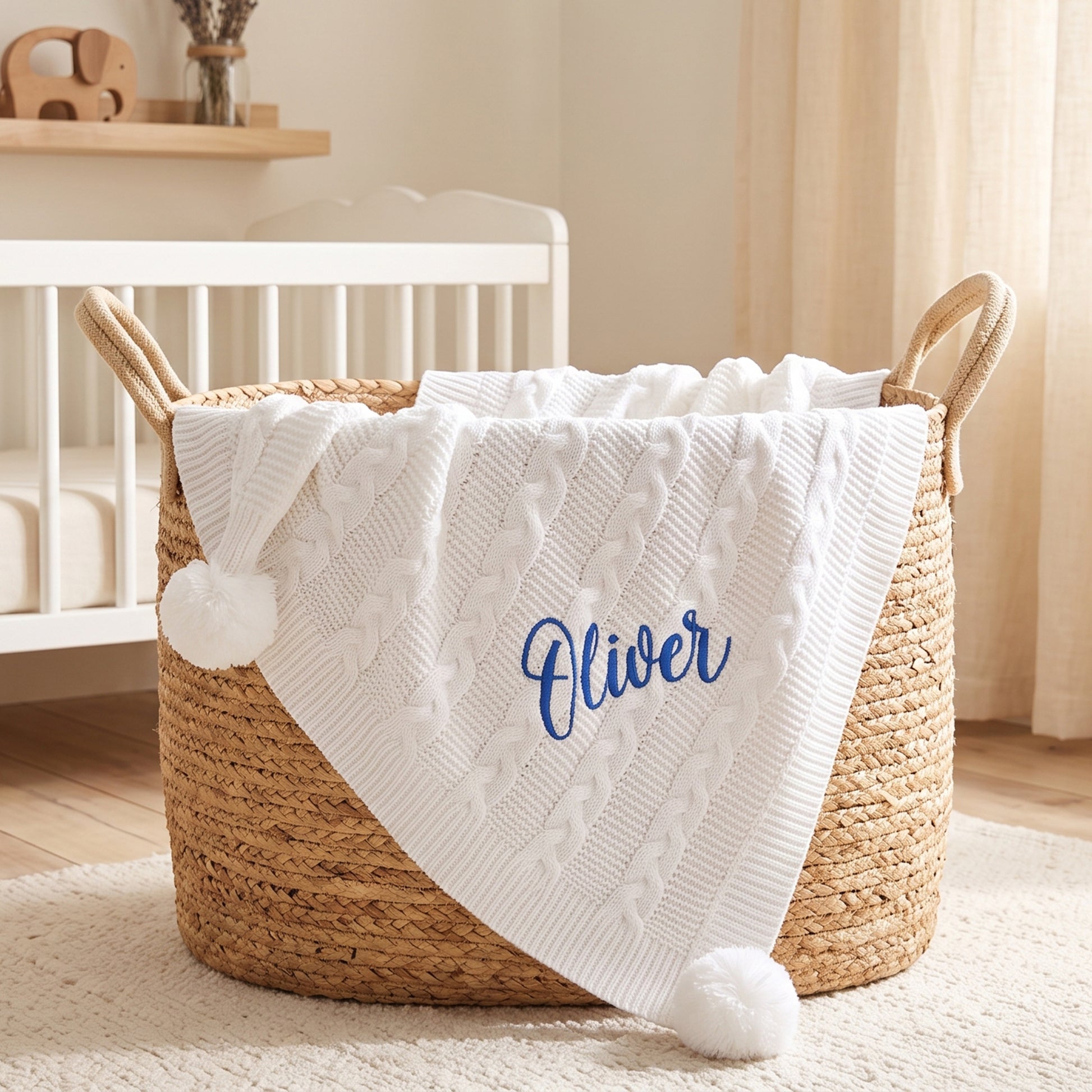 White cable knit baby blanket with pom-poms at each corner, personalised with the name Oliver in blue embroidery, draped over a woven basket.