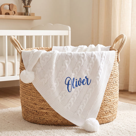 White cable knit baby blanket with pom-poms at each corner, personalised with the name Oliver in blue embroidery, draped over a woven basket.