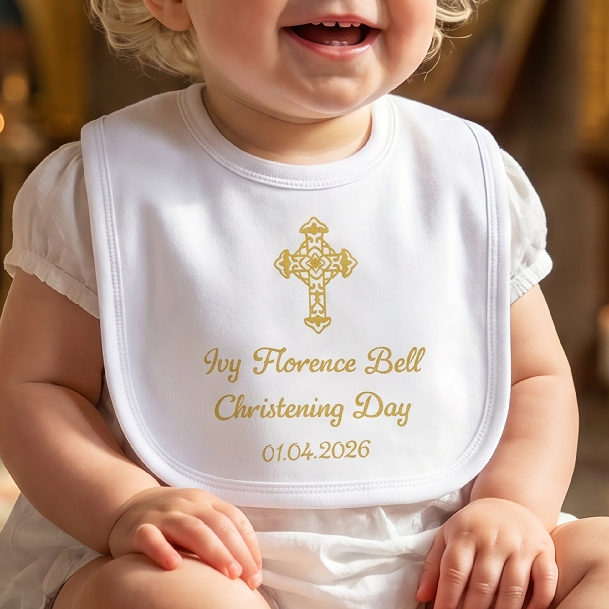 White velcro christening bib printed with a gold cross and personalised with the name Ivy Florence Bell, "Christening Day" and the date 01.04.2026, worn by a smiling baby.