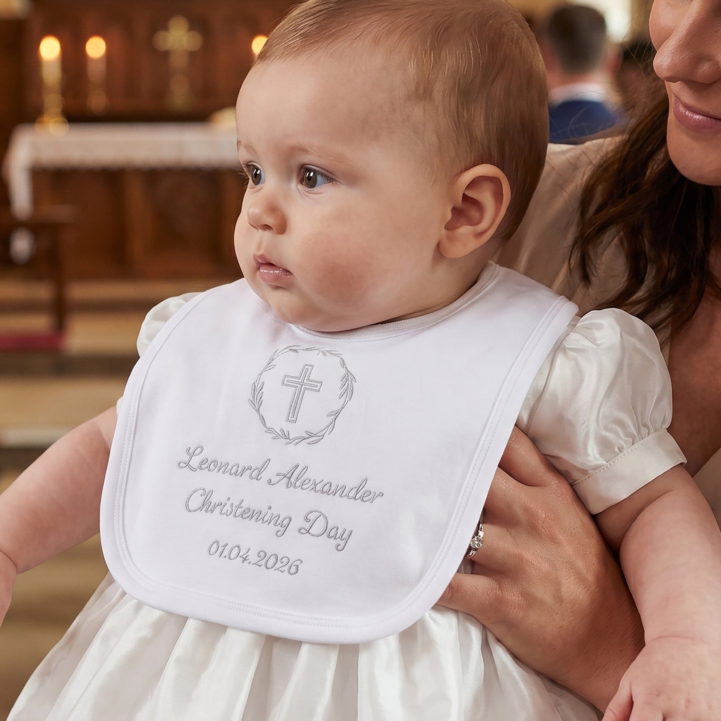 White velcro christening bib embroidered with a grey cross inside a laurel wreath, personalised with the name Leonard Alexander, "Christening Day" and the date 01.04.2026, worn by a baby held by a parent.
