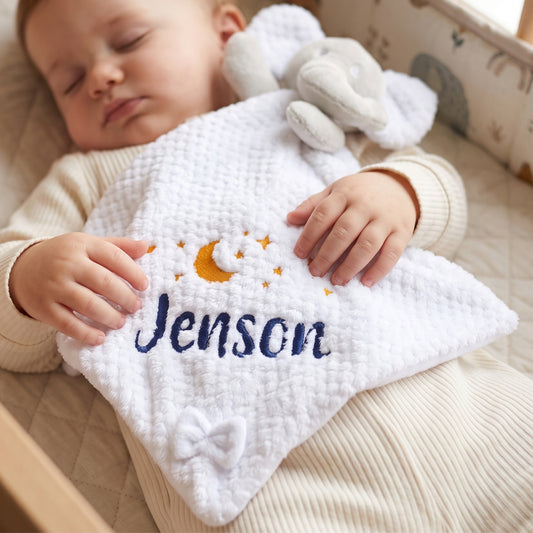 A sleeping baby lying in a crib, holding a white textured comforter with an elephant, moon, stars, and the name 'Jenson' embroidered in blue and yellow.