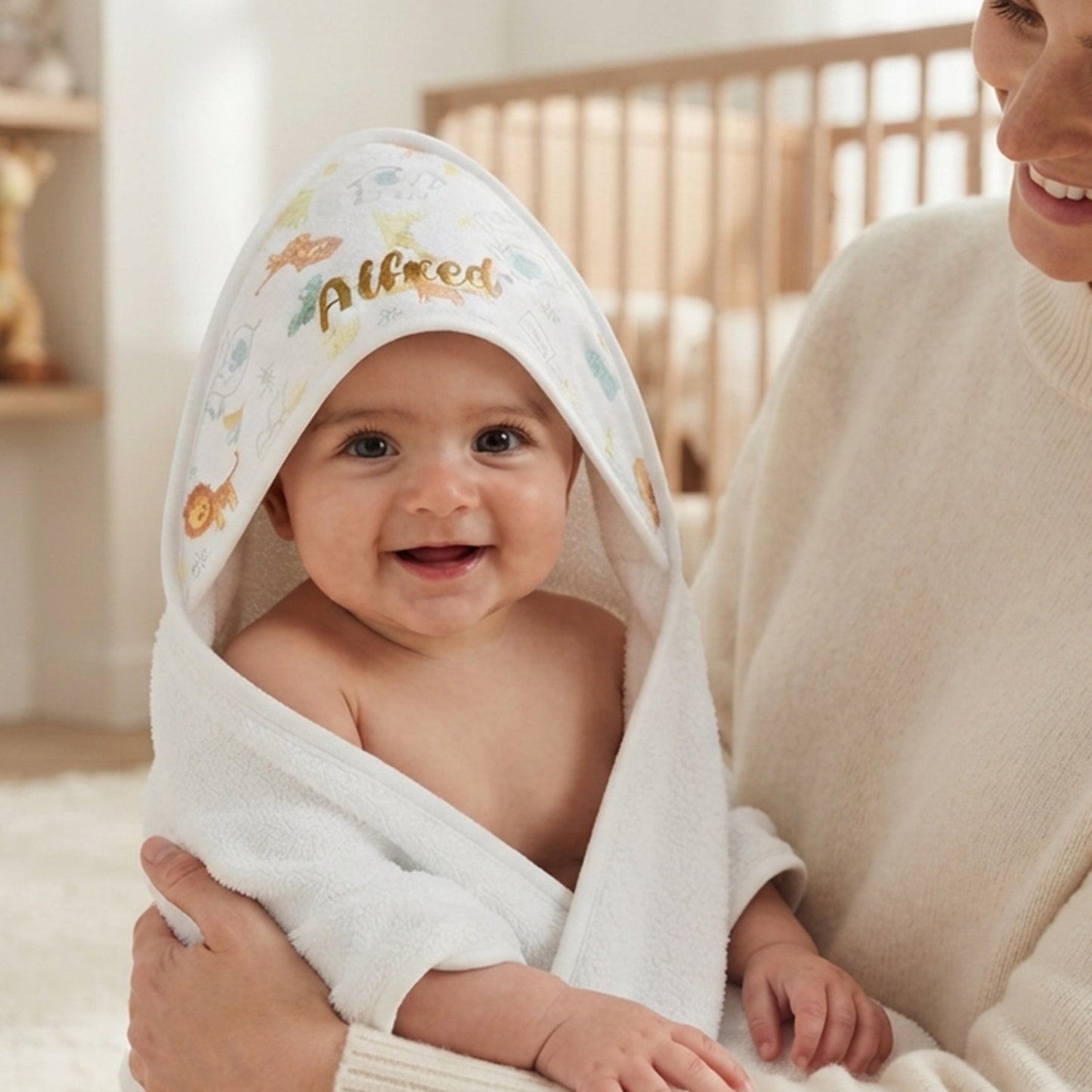 A baby wrapped in a white hooded towel with colorful animal illustrations and the name 'Alfred' embroidered on the hood, held by an adult in a cream sweater in a softly lit indoor setting.