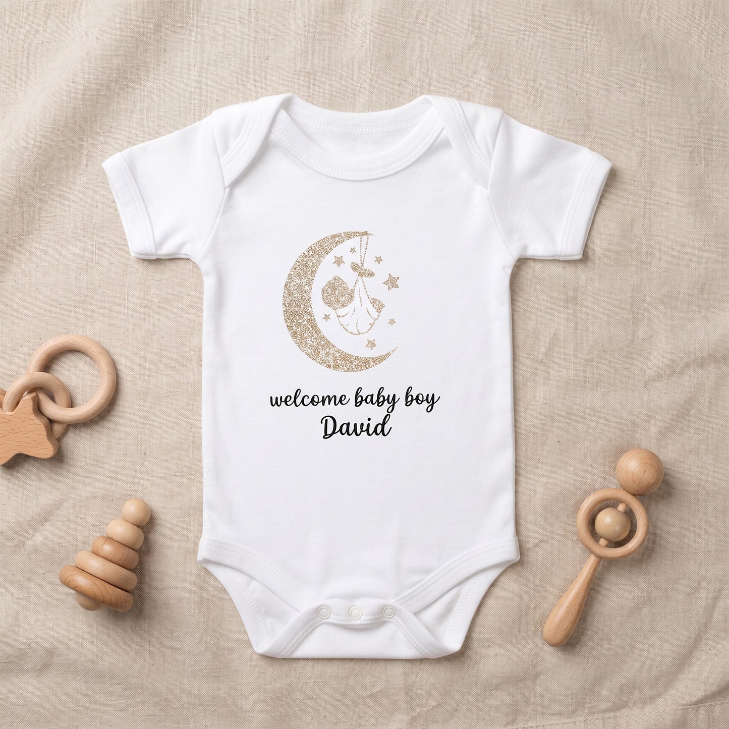 A white short-sleeve baby bodysuit laid flat on a beige fabric background, printed with a gold glitter crescent moon and stars graphic and the words 'welcome baby boy David' in black. Wooden baby toys are placed around the bodysuit.