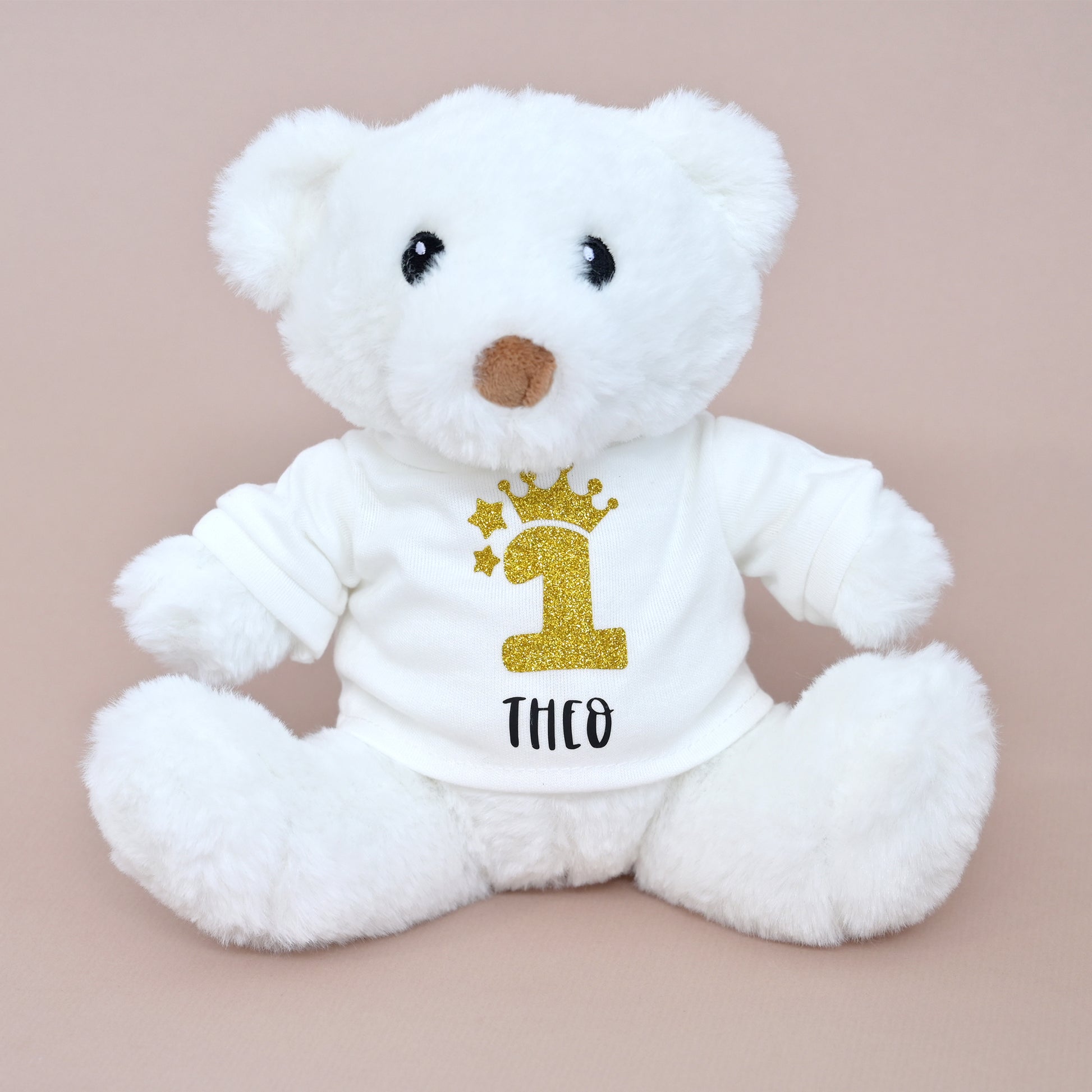 A white plush teddy bear wearing a white long-sleeve shirt with a glittery gold number 1 and the name 'THCO' printed in black, set against a solid pale pink background.