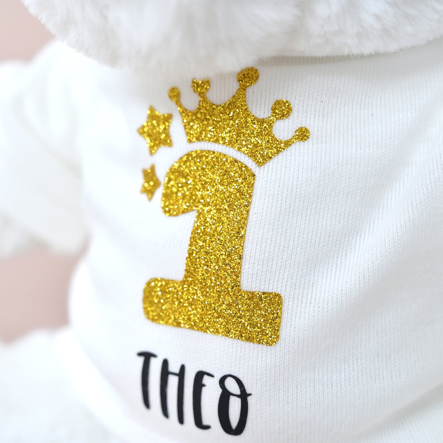 A close-up of a white pet shirt with a glittery gold number 1 wearing a crown and the name 'THEO' printed below in black.