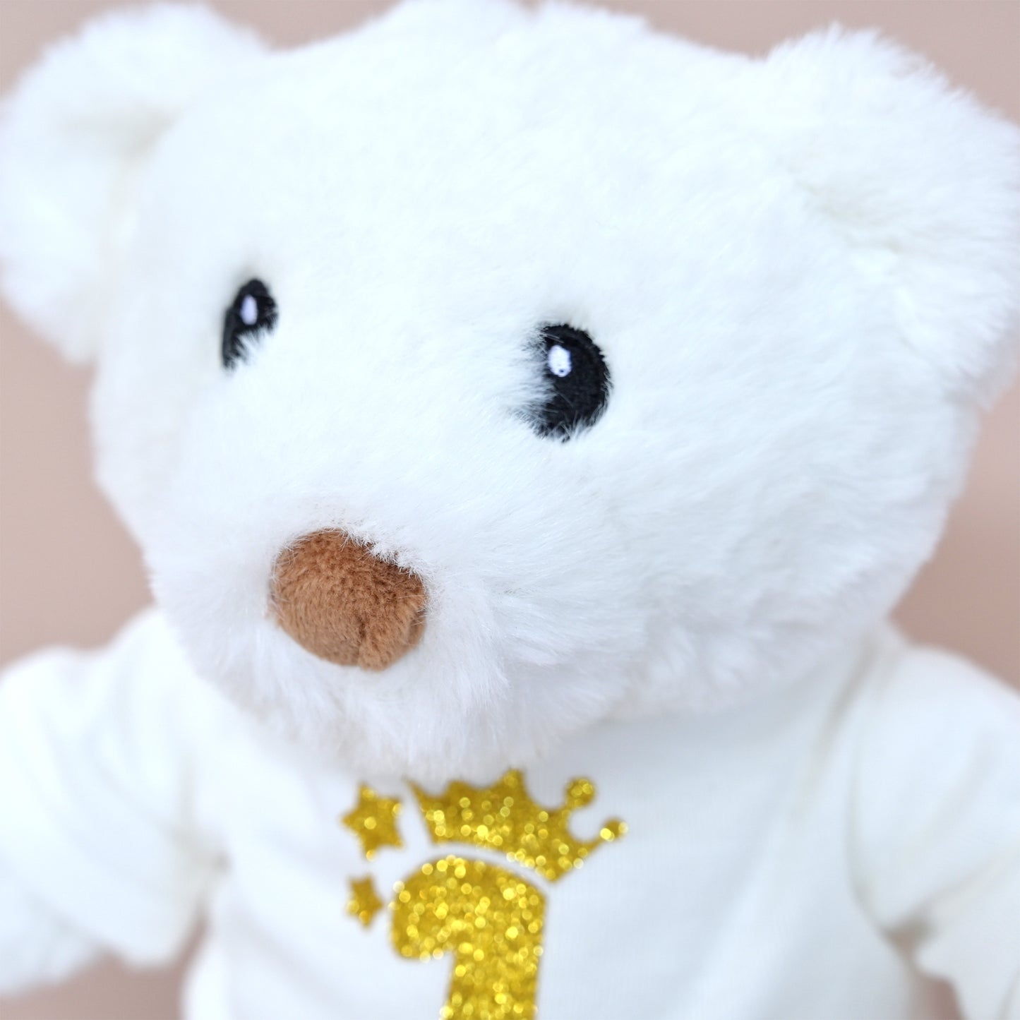 A close-up of a white plush teddy bear wearing a white shirt with a glittery gold number 1 and crown design on the chest, set against a soft beige background.