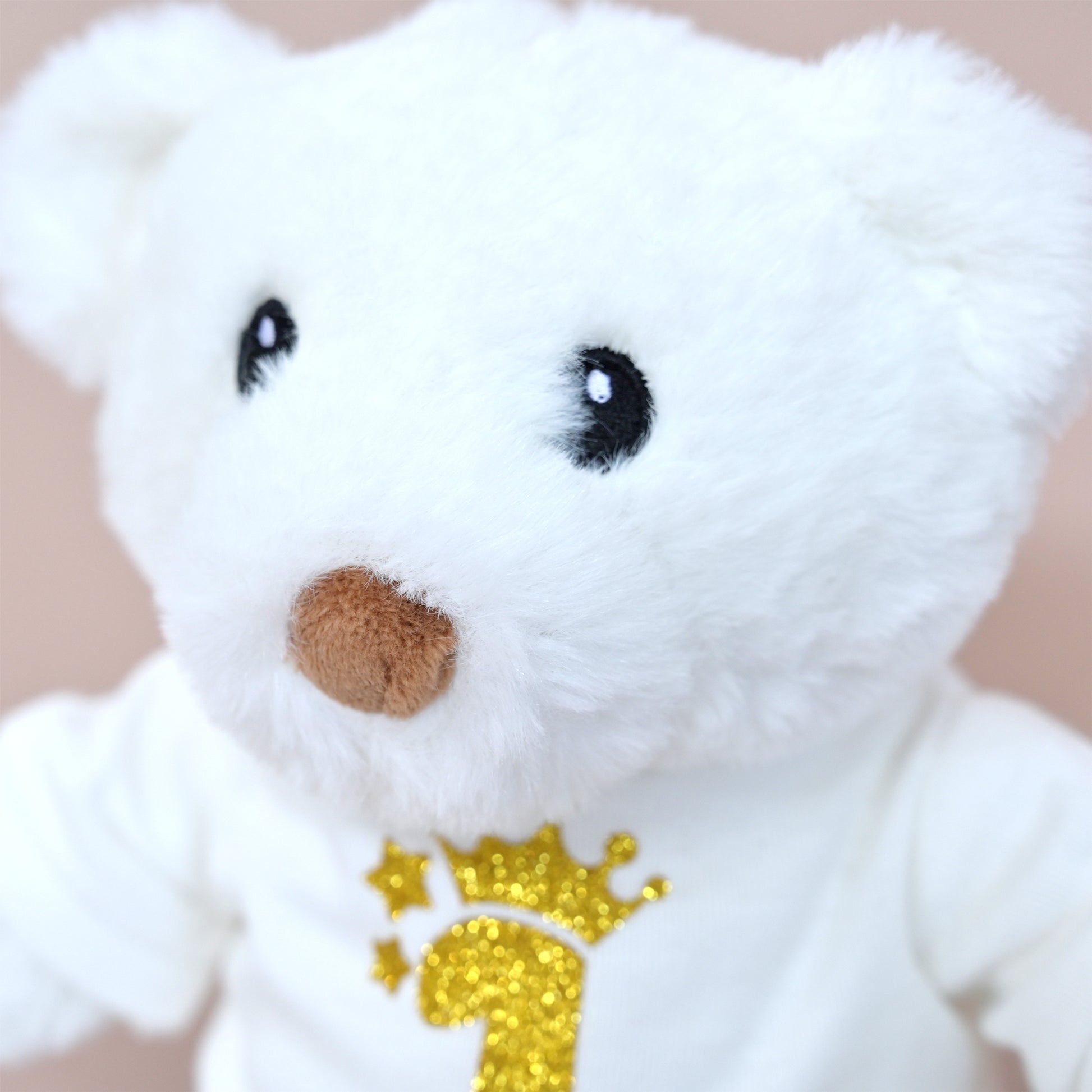 A close-up of a white plush teddy bear wearing a white shirt with a glittery gold number 1 and crown design on the chest, set against a soft beige background.