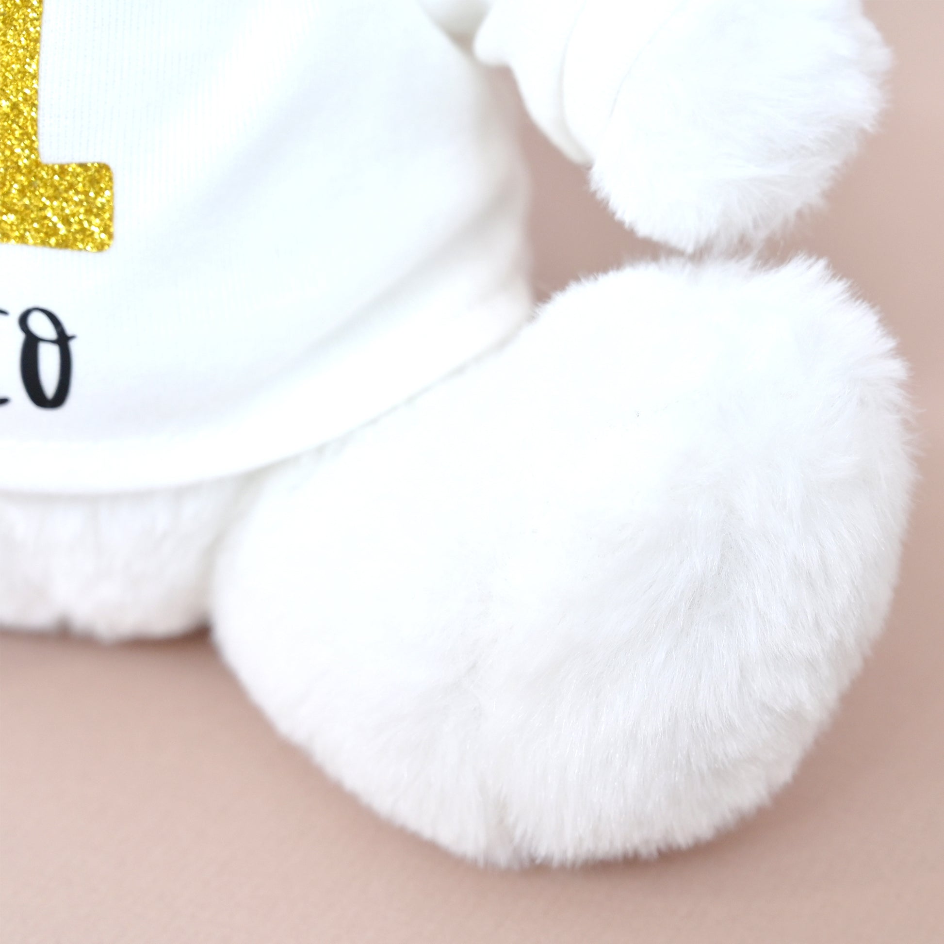 Close-up of a white plush toy or stuffed animal wearing a white shirt with a glittery gold letter and black text, resting on a soft beige surface.
