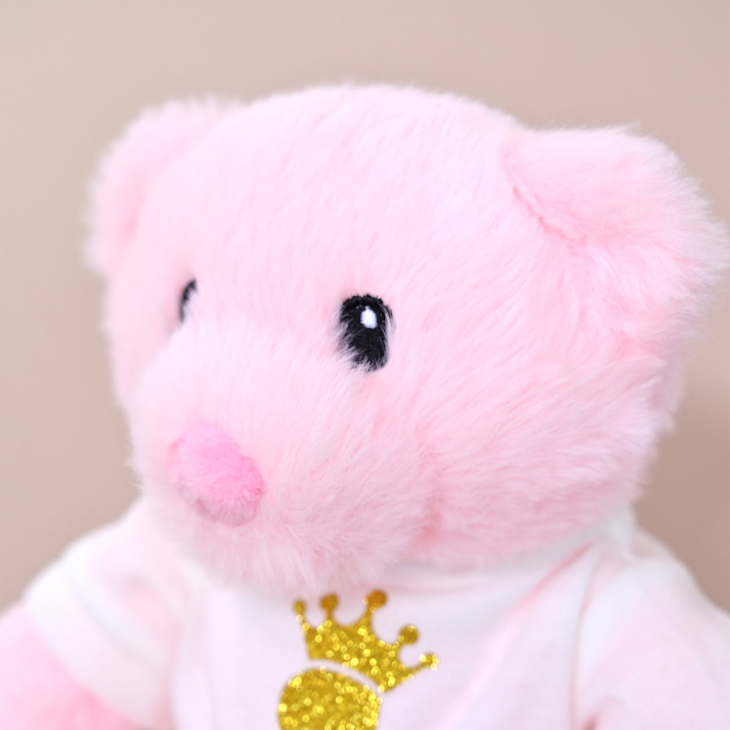 A close-up of a soft pink plush pig toy wearing a white shirt with a glittery gold crown graphic, set against a plain beige background.