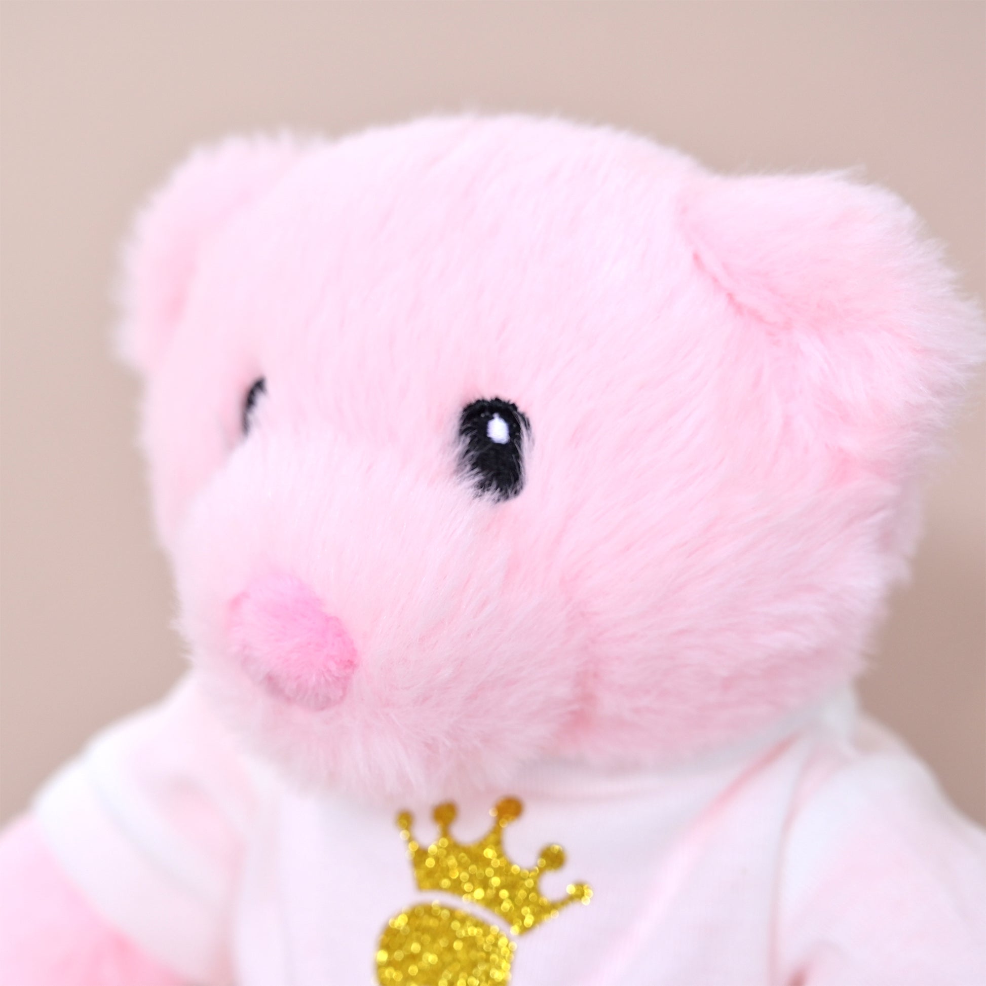 A close-up of a soft pink plush pig toy wearing a white shirt with a glittery gold crown graphic, set against a plain beige background.