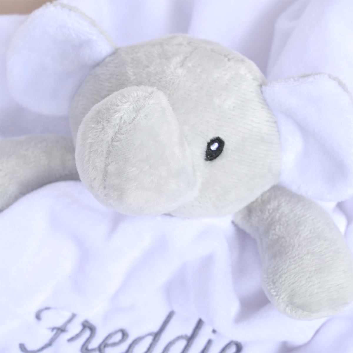 A close-up of a plush elephant comforter or blanket in light gray, resting on a white fabric with the name 'Freddie' embroidered in gray script.