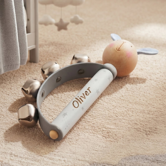 A personalized baby sensory toy with a wooden rabbit head, silver bells, and a white handle engraved with the name 'Oliver', resting on a beige carpet.