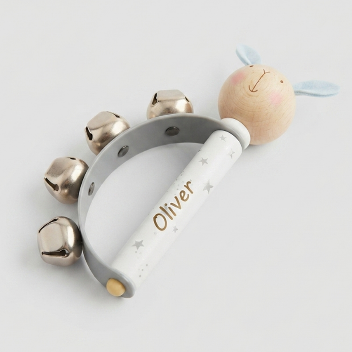 Personalized wooden baby rattle with five gold bells, a white handle printed with the name 'Oliver' and small stars, and a light wood bunny head with blue ears on a light gray background.