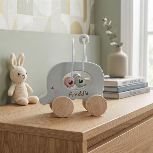 A personalized wooden toy car shaped like an elephant, with the name 'Freddie' written on the side, displayed on a wooden dresser in a styled nursery setting next to a knitted bunny toy.