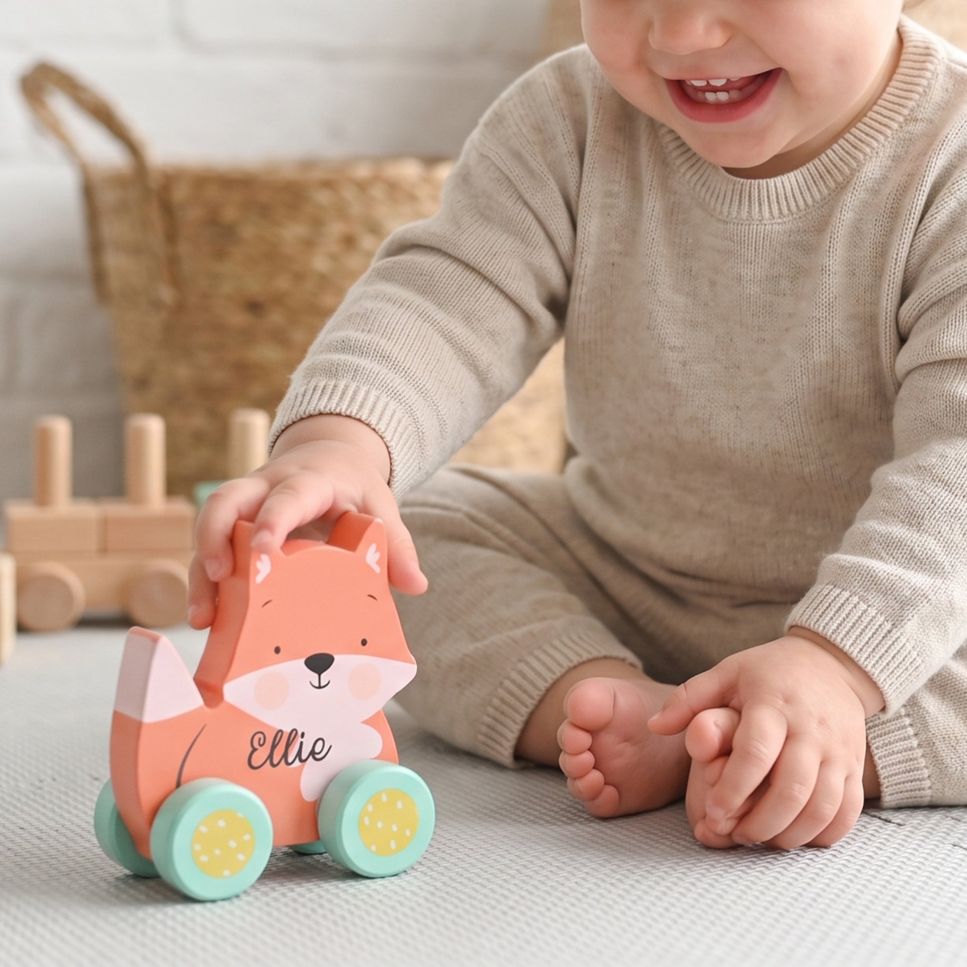 A baby in a beige knit outfit plays with a wooden fox toy car on a light textured floor. The toy is orange-pink with mint green wheels and the name 'Ellie' printed on the side.