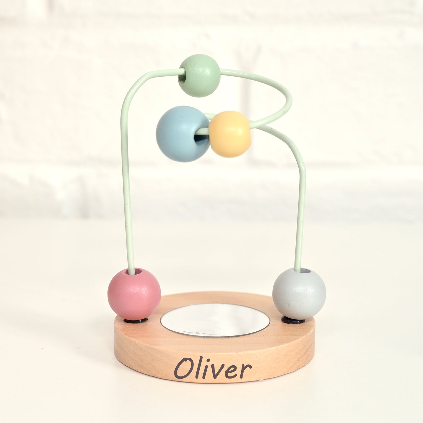 A personalized wooden bead maze toy for children, featuring a light natural wood base engraved with the name 'Oliver', a mint green metal frame, and four pastel-colored wooden beads (pink, light blue, yellow, and light gray).