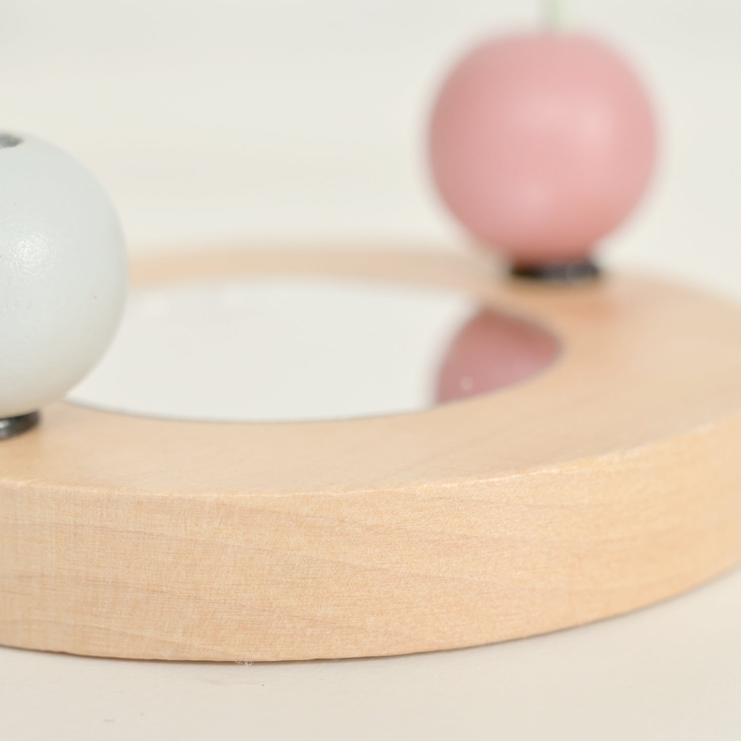 A close-up of a light natural wood circular tray or dish with a shallow center recess, featuring a white spherical object on the left and a soft pink spherical object on the right, set against a warm off-white background.