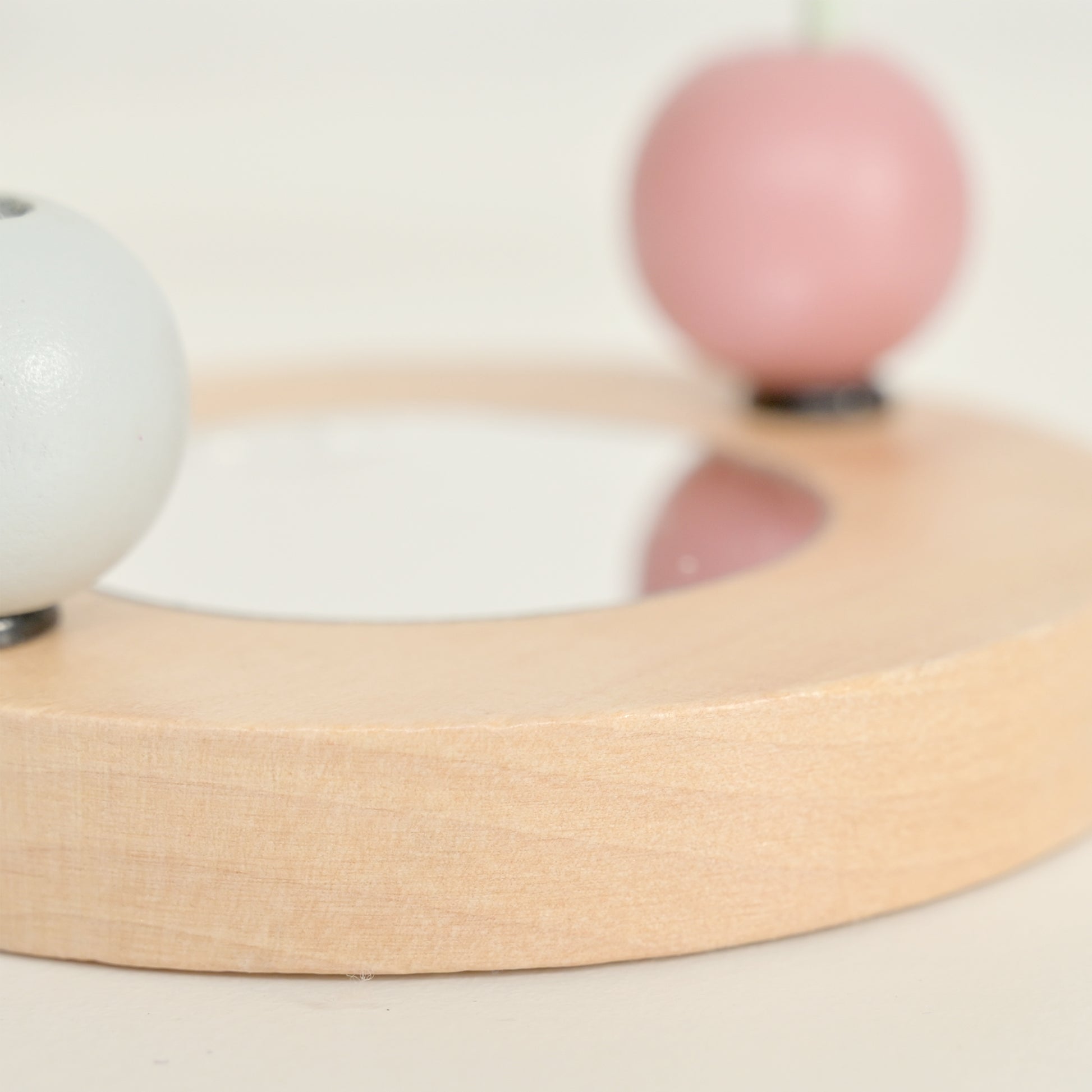 A close-up of a light natural wood circular tray or dish with a shallow center recess, featuring a white spherical object on the left and a soft pink spherical object on the right, set against a warm off-white background.