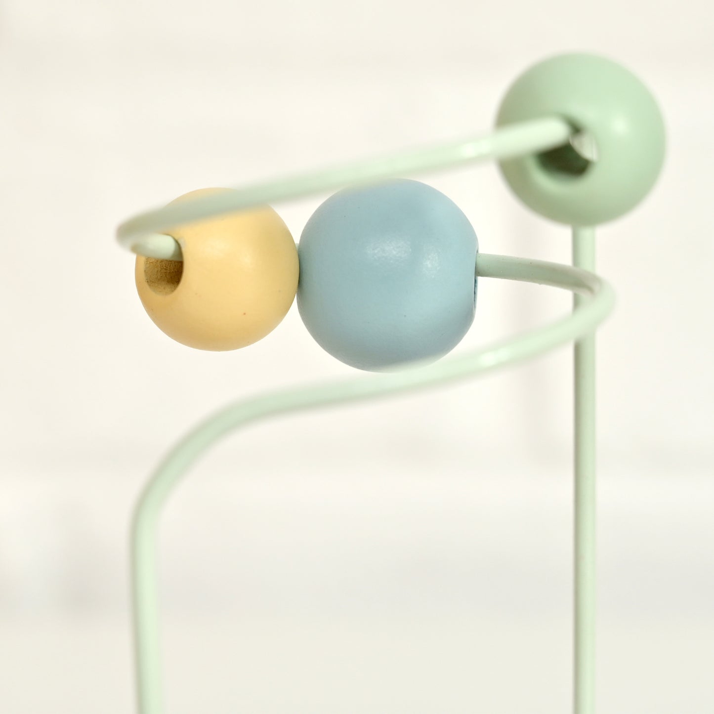 A close-up of a pastel-colored wooden bead maze toy with three round beads in mint green, light blue, and pale yellow on a mint green metal frame, set against a soft white background.
