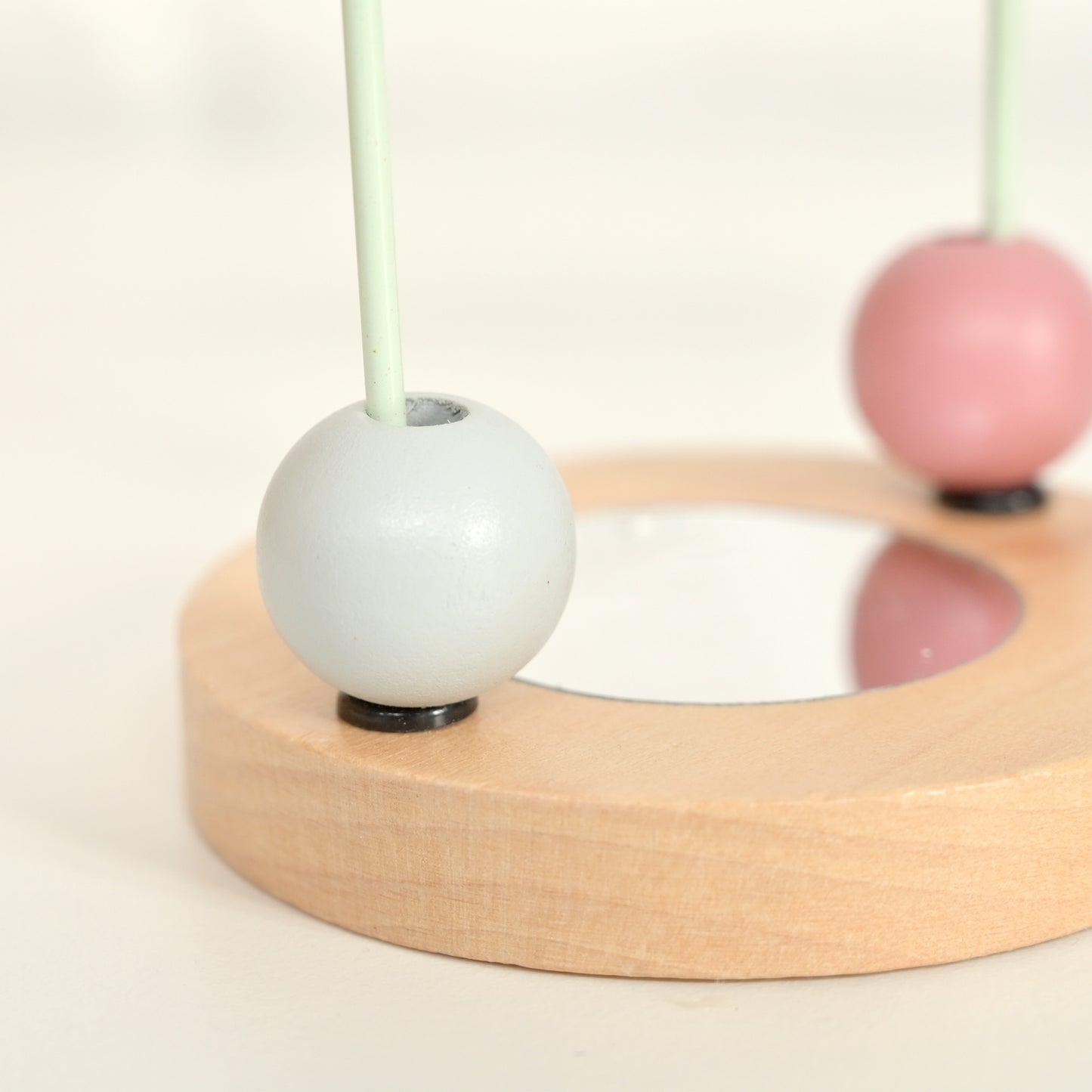 A close-up of a wooden bead activity board with a light natural wood base, a small mirror in the center, a pale gray bead in the foreground, and a pink bead in the background.