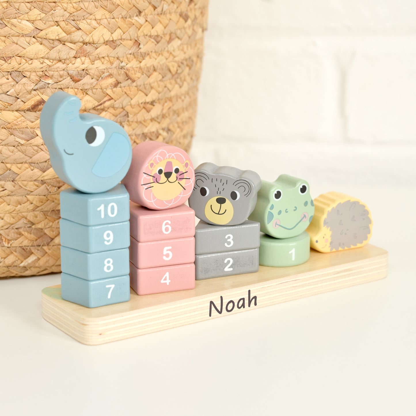 Personalized wooden number blocks and animal-shaped stacking pieces on a light wood base labeled 'Noah', photographed on a white surface with a woven basket and white brick wall in the background.
