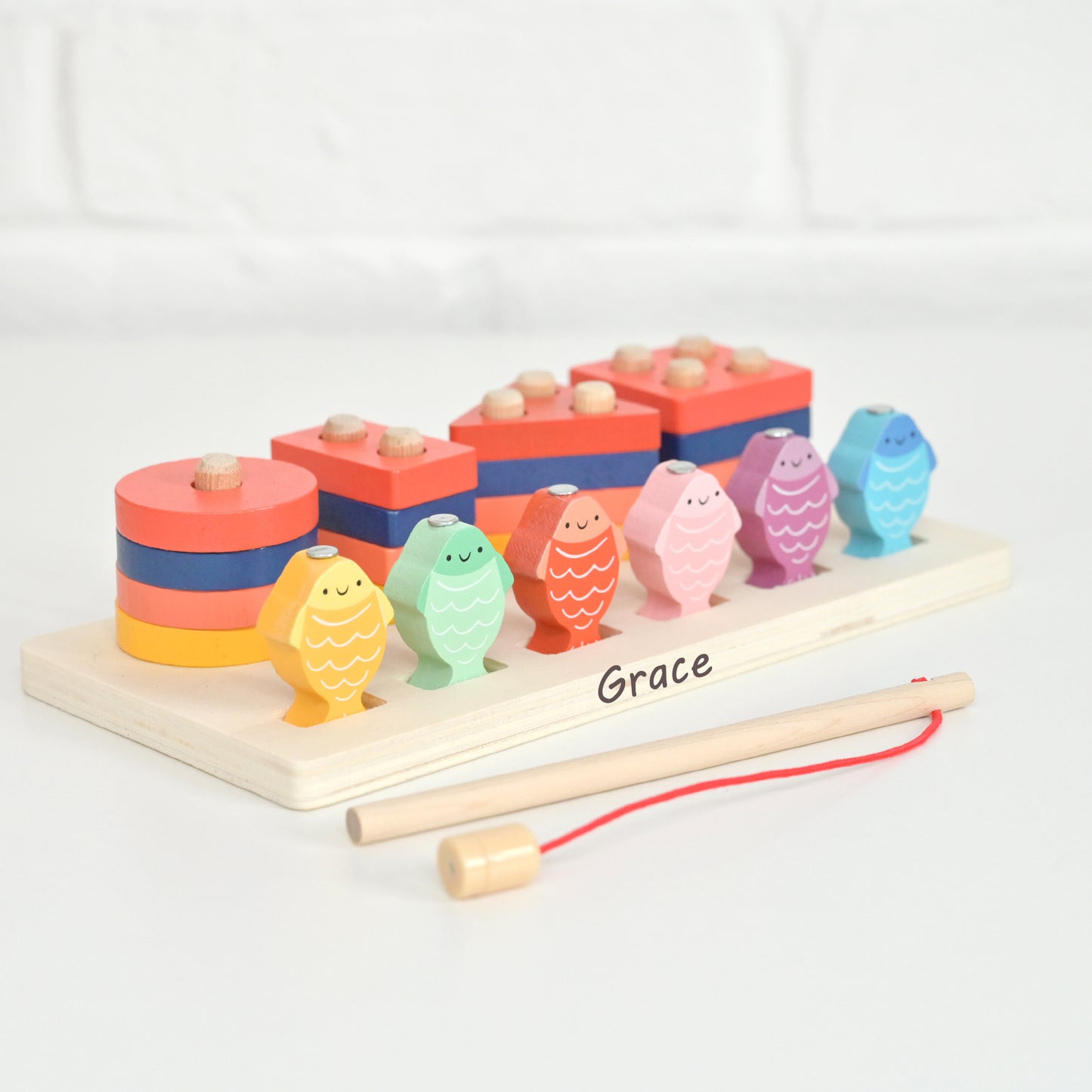 A personalized wooden toy block set with colorful fish-shaped pegs and stacking blocks on a light wood base, labeled 'Grace', with two wooden mallets and a red string in front, photographed on a white surface against a white brick wall.