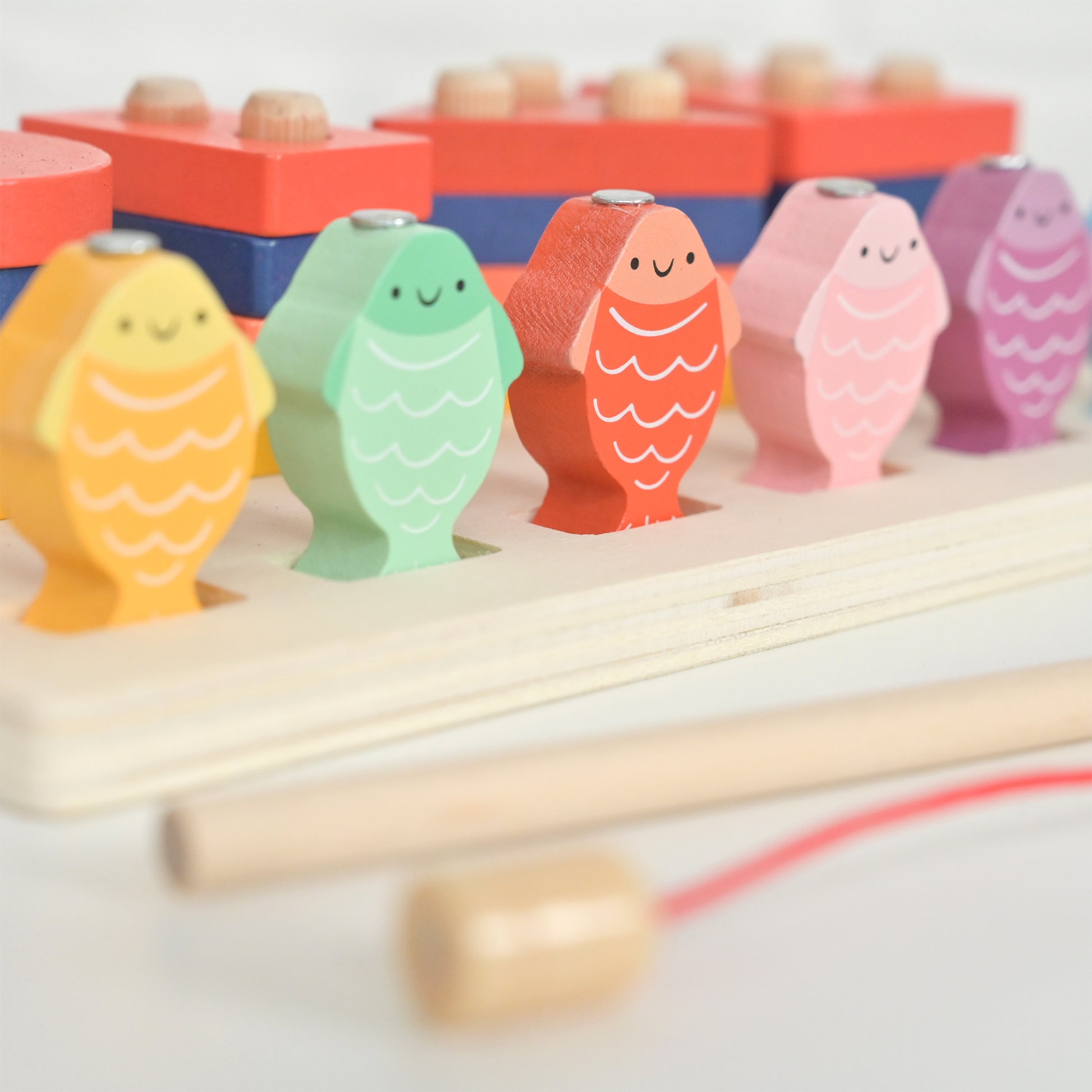 A colorful wooden fish toy with six smiling fish-shaped pegs in yellow, mint green, coral orange, pink, and purple, set on a light wood base with a wooden mallet and red string.