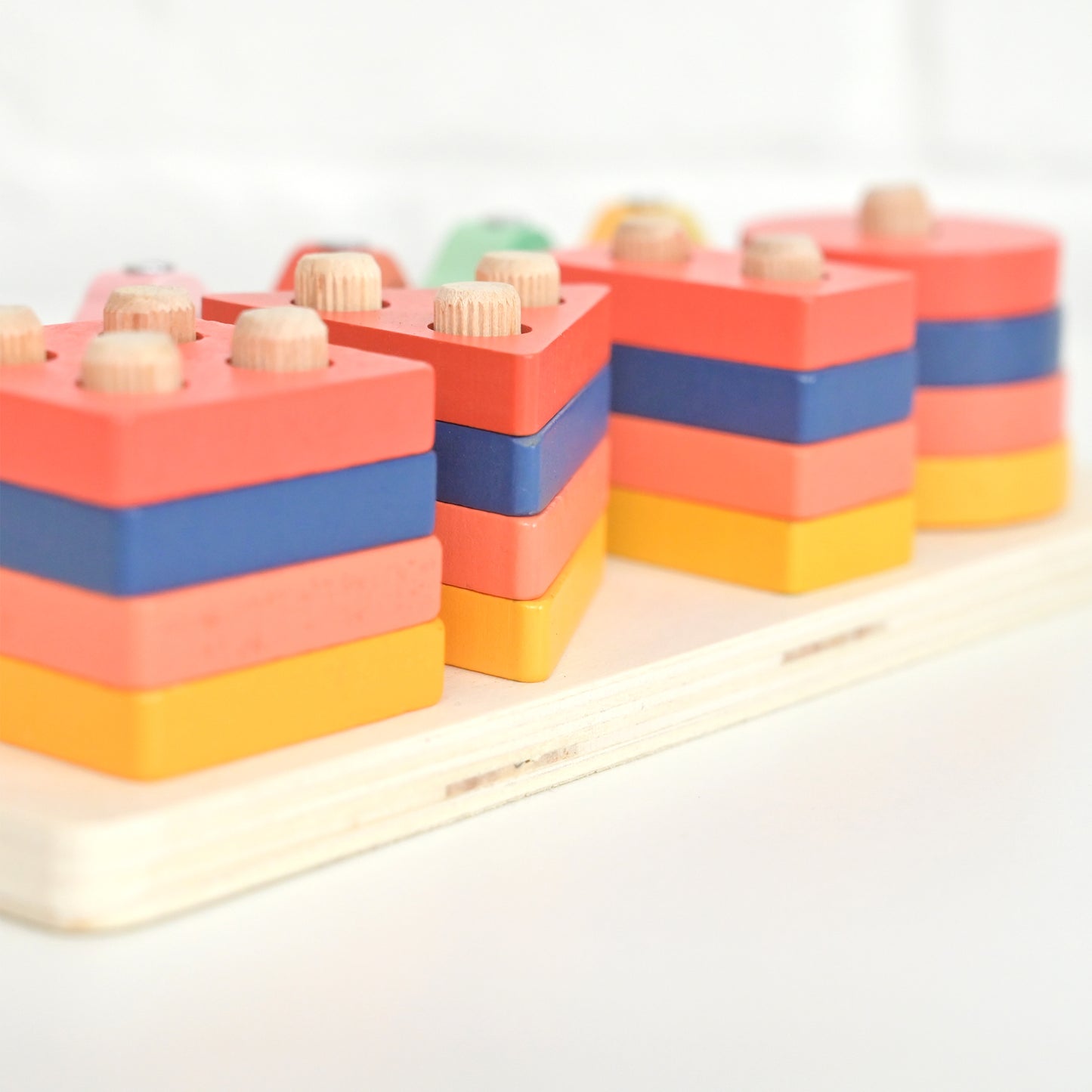 A set of colorful wooden stacking blocks arranged on a light wood tray. The blocks are made of layered wood in coral pink, royal blue, and yellow, with small natural wood pegs on top, photographed against a bright white background.