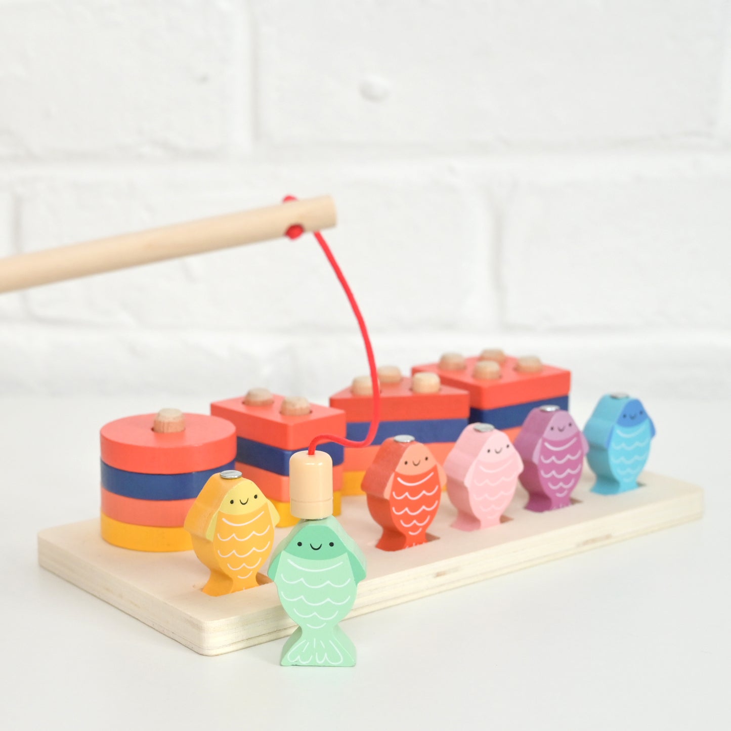 A colorful wooden toy fishing set on a light natural wood base, featuring fish-shaped hooks in pastel and bright colors, stacked wooden blocks, and a wooden fishing rod with a red string, photographed against a white brick wall background.