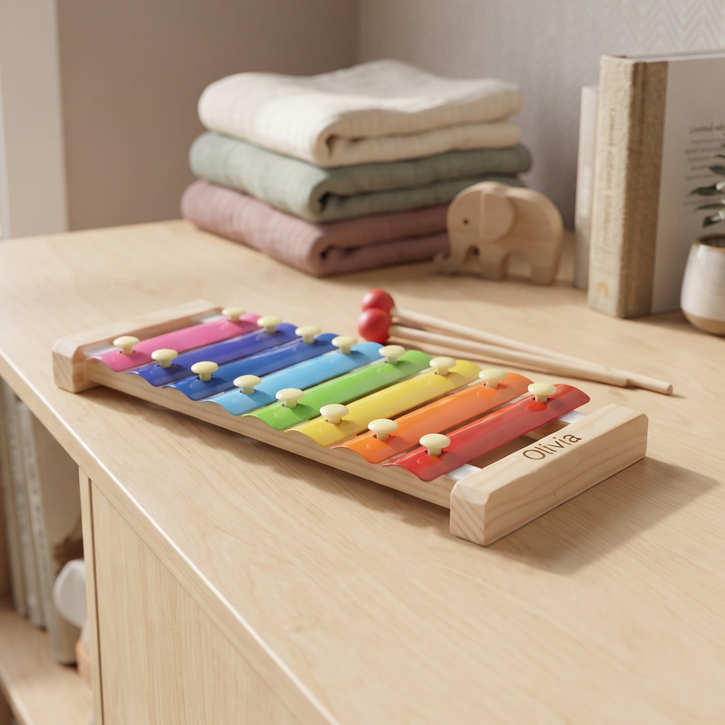 A wooden xylophone toy with colorful painted keys and two wooden mallets, placed on a light wood table in a softly styled room. The xylophone has the name 'Olivia' engraved on the side.