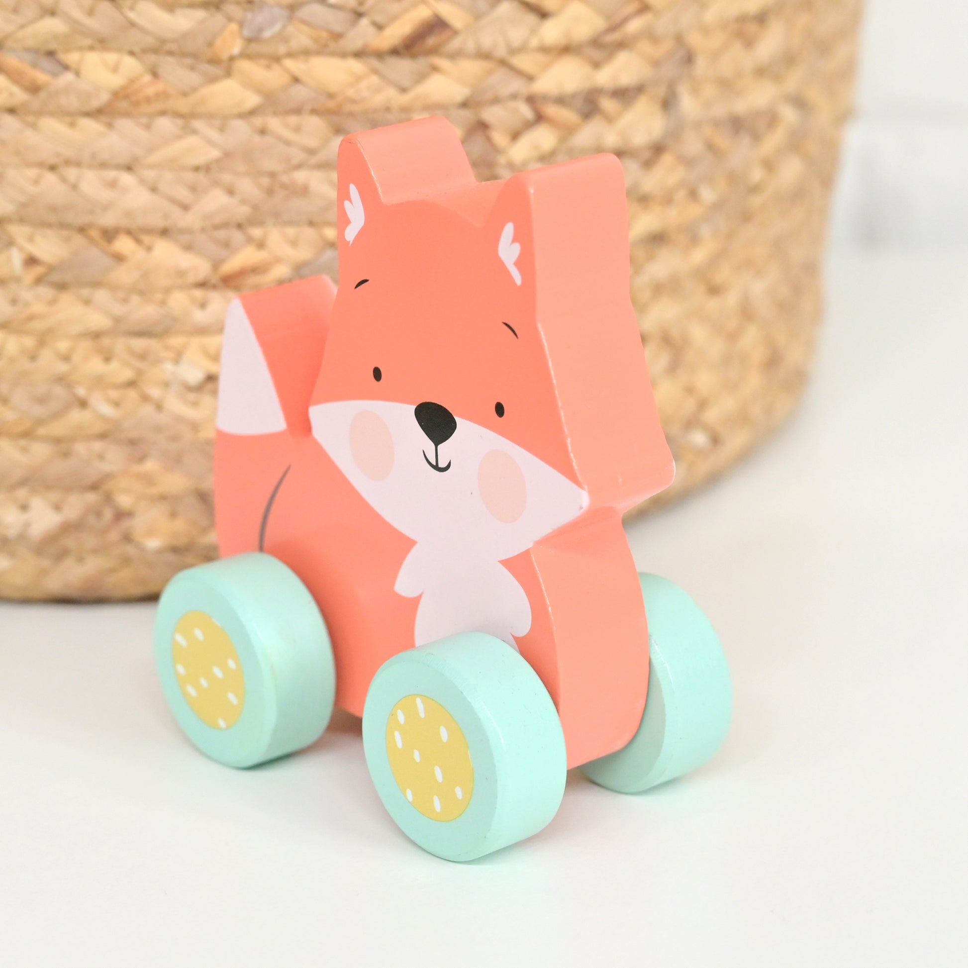 A wooden fox toy car with a coral-orange body, white face, and mint green wheels with yellow centers, photographed on a white surface with a woven basket in the background.