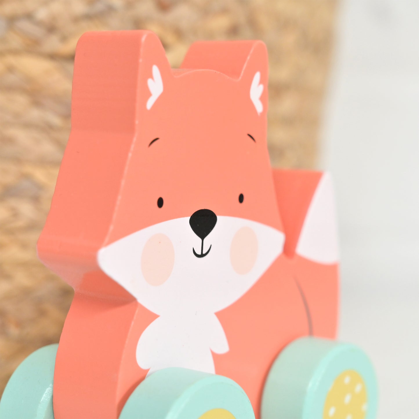 A close-up of a wooden toy fox with a coral-orange body, white face and chest, black nose and eyes, and mint green wheels with yellow details, photographed against a softly blurred neutral background.