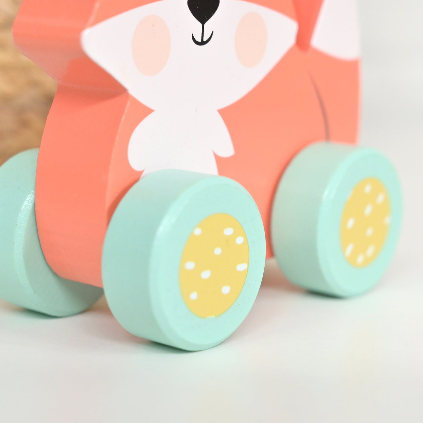 A close-up of a wooden toy cart with a smiling fox face on the front, featuring a coral-orange body, white face, mint green wheels, and yellow wheel centers with white dots.