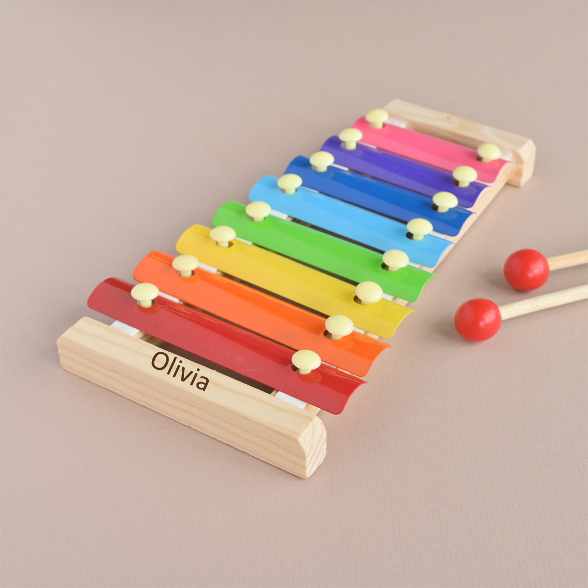 A personalized wooden xylophone toy with rainbow-colored metal bars, engraved with the name 'Olivia' on the red bar, shown with two red mallets on a neutral beige background.
