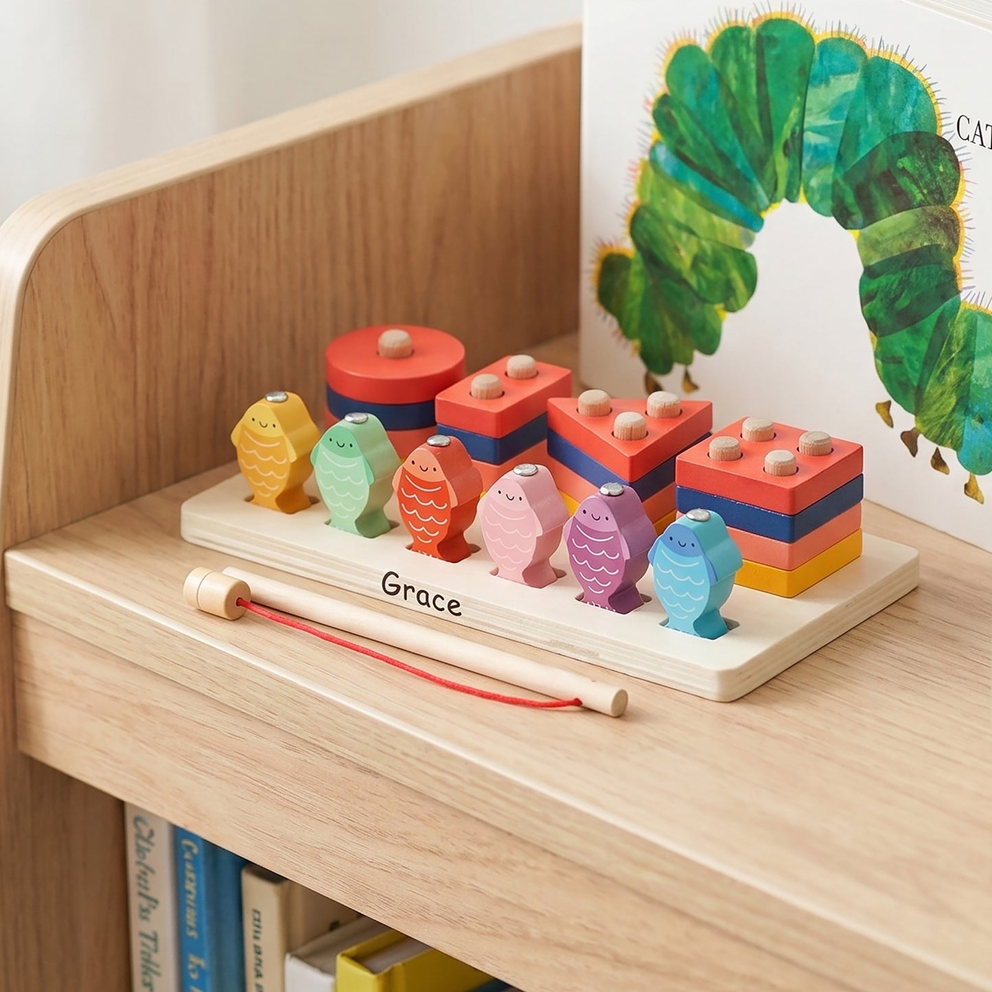 A colorful wooden children's toy set on a light wood shelf, featuring a base labeled 'Grace' with fish-shaped pegs, stacked block towers, and a wooden pick with a red string. A children's book with a green caterpillar illustration is partially visible in the background.
